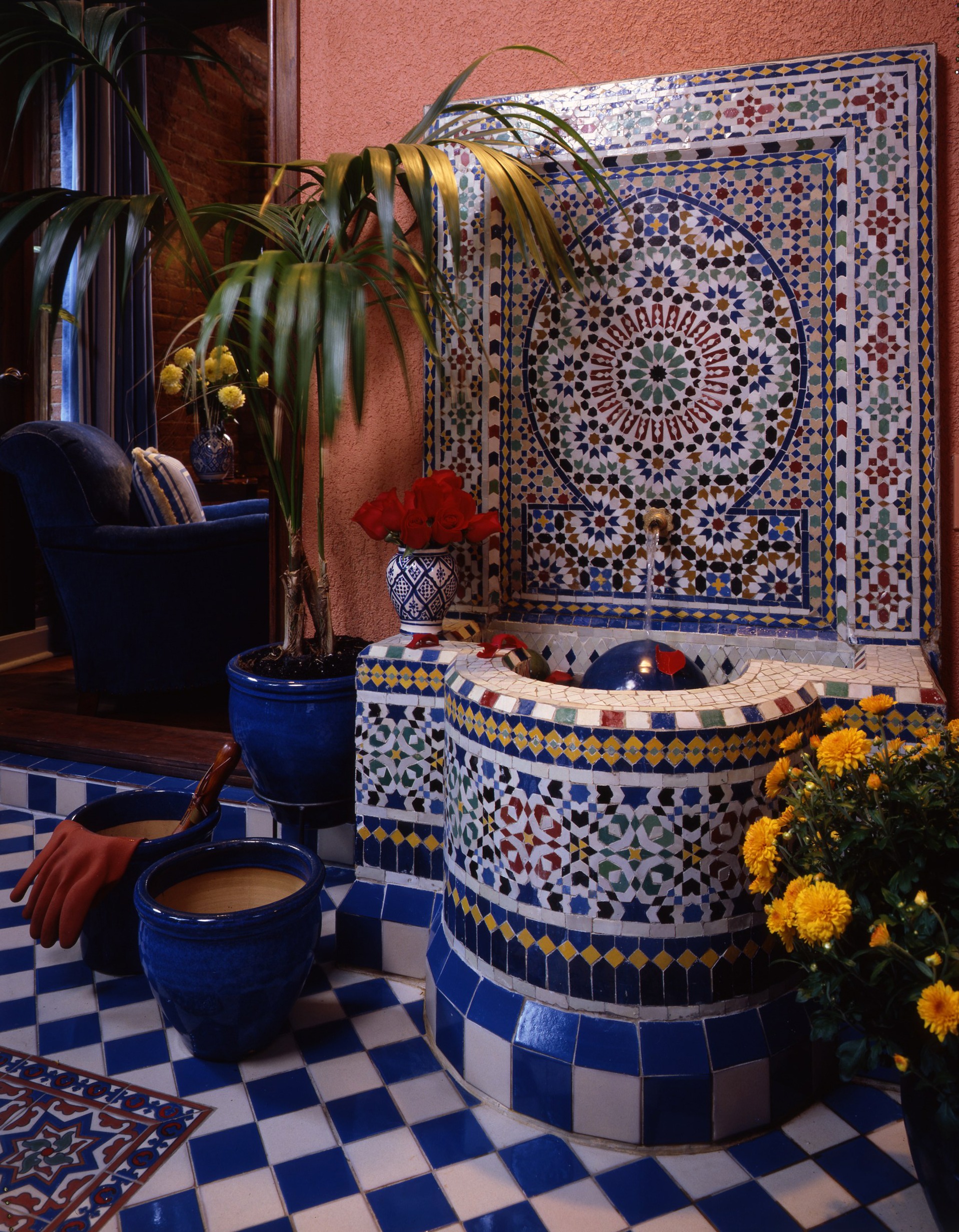 Colorful tiled fountain with intricate patterns, surrounded by potted plants and flowers, creates a serene indoor space. Ideal for relaxation and aesthetic appeal.