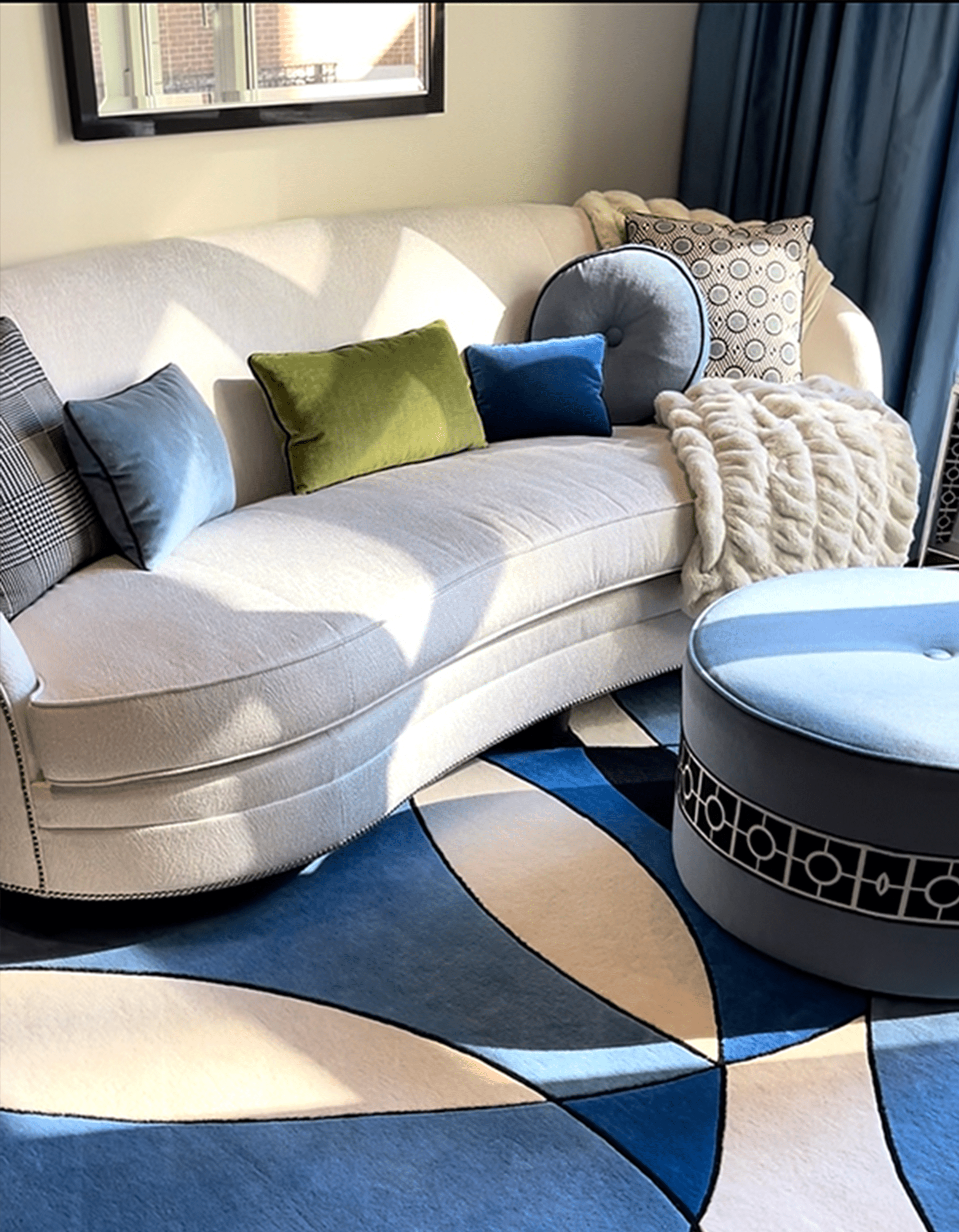 Curved white sofa accented with colorful pillows, draped with a throw, beside a round blue ottoman on a patterned rug featuring blue and cream shapes.